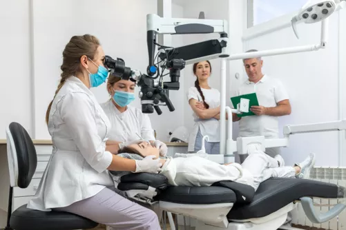 a dentist looking through a microscope over a patient with other dentists standing around with masks on 